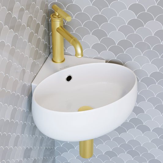 Small white wall-hung corner sink with single hole and gold matte faucet against textured gray tile wall.